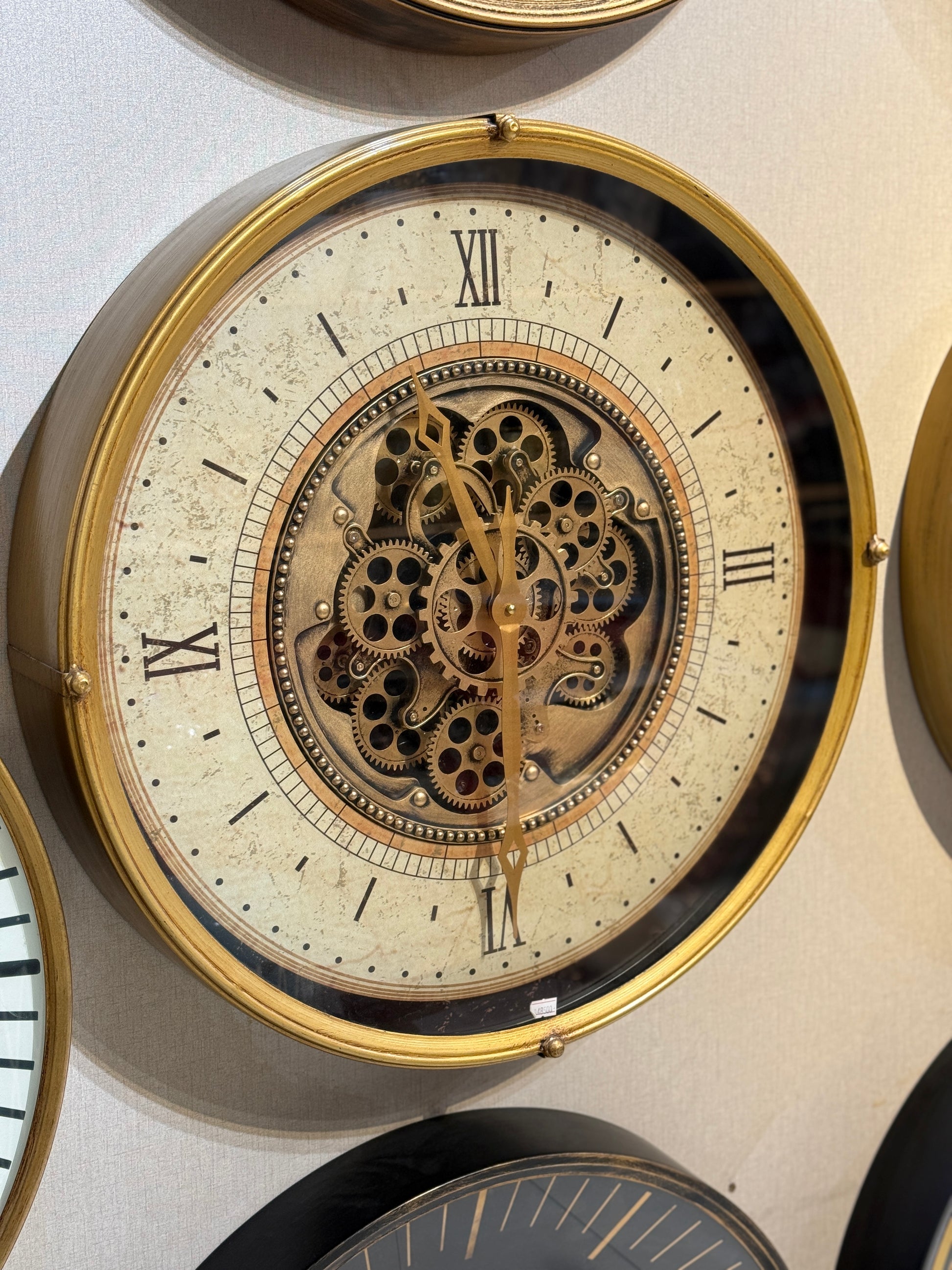 Large 23-inch diameter gold metal wall clock with visible mechanical gears and Roman numerals at Fatima Home Décor store in Islamabad.