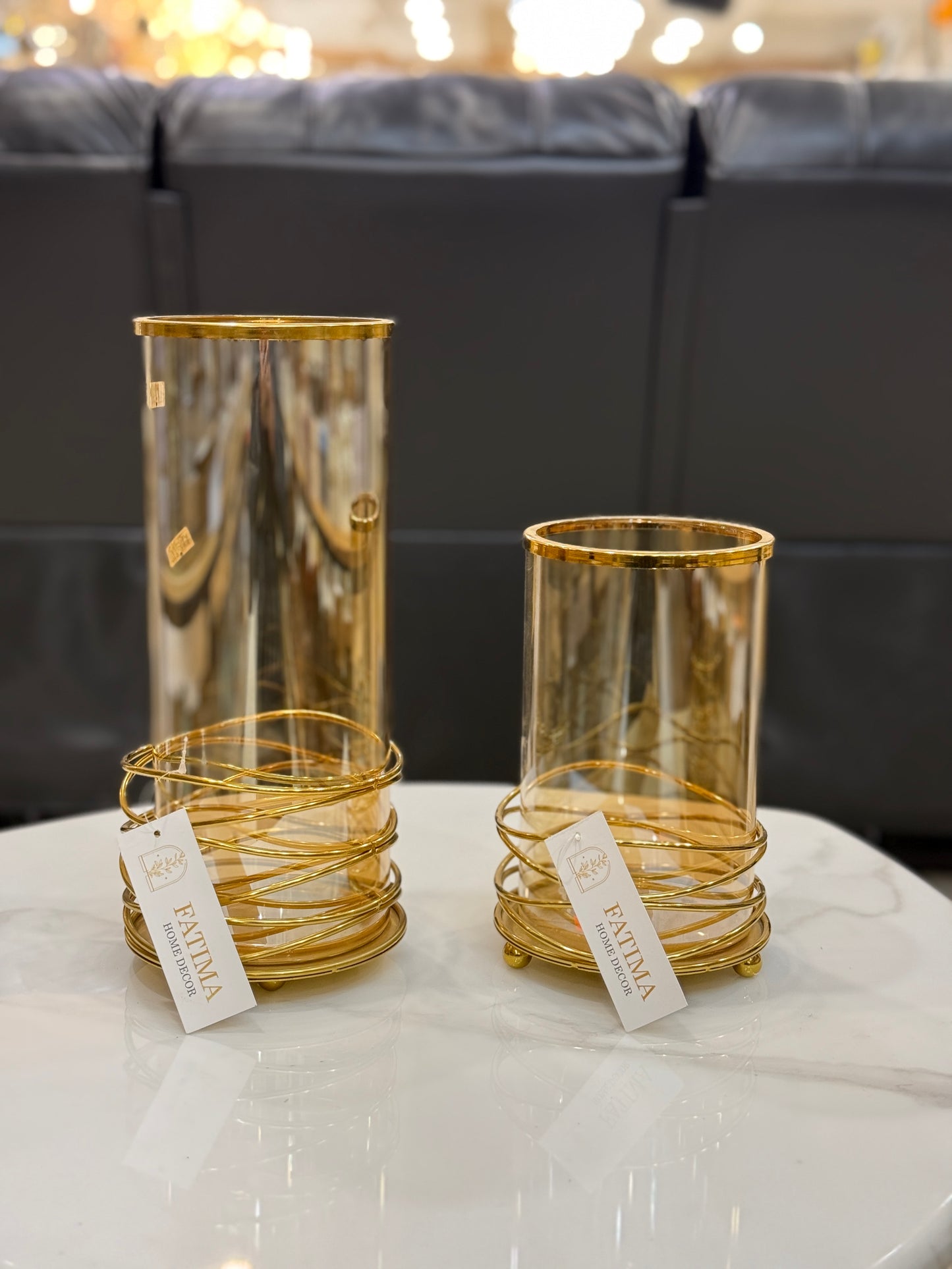 Pair of amber glass hurricane candle holders with gold rims and golden wire bases on a white marble table at Fatima Home Decor store.