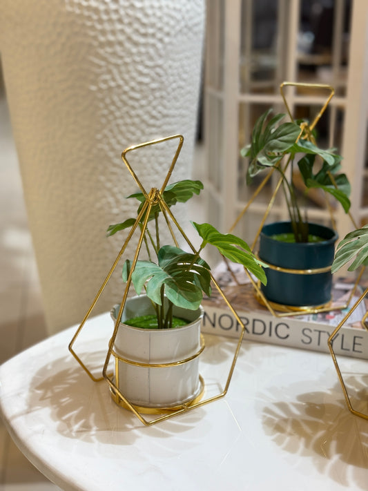 Artificial Mons tera plant in a teal pot inside a gold triangular geometric frame displayed on a Nordic Style book at Fatima Home Décor Islamabad.
