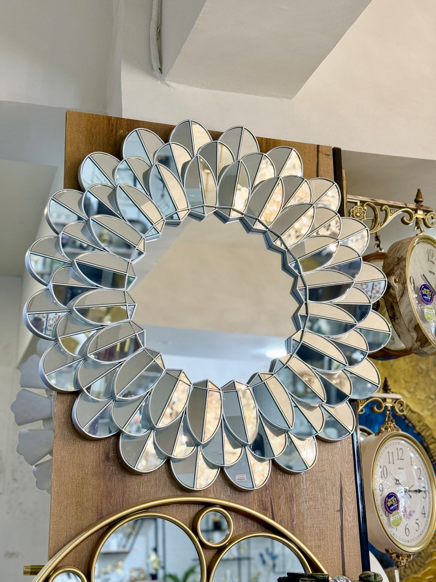 Silver petal-shaped decorative wall mirror with PVC frame displayed on a wooden wall at Fatima Home Decor store in Islamabad.
